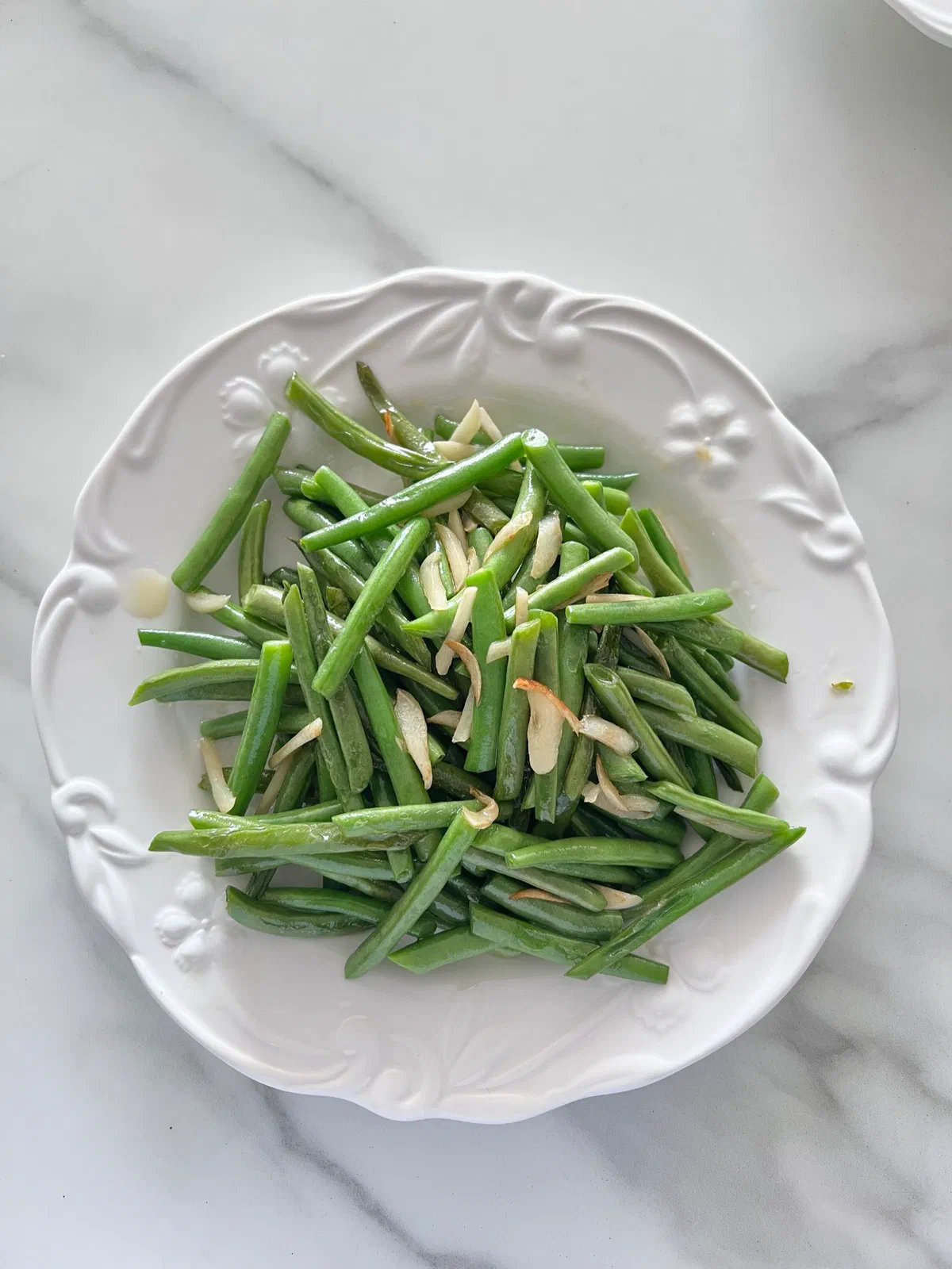 Une assiette blanche de haricots verts cuits parsemés d'amandes effilées, sur un plan en marbre