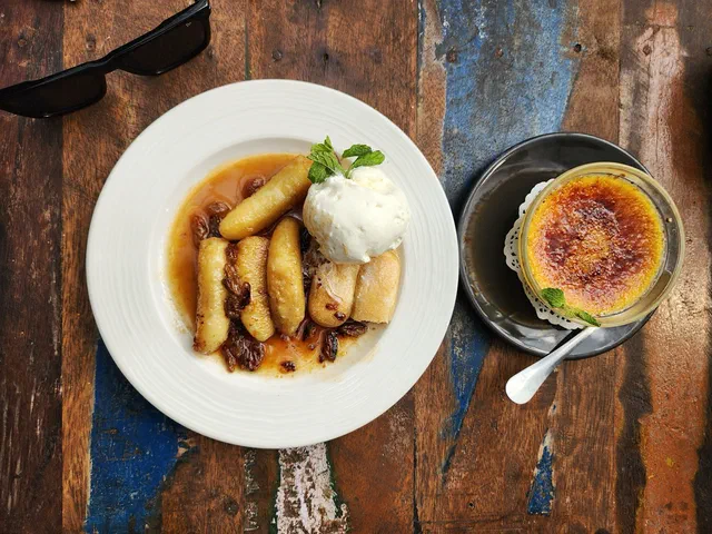 Une assiette de bananes caramélisées dans leur sauce avec une boule de glace vanille, à côté d'une crème brûlée, sur une table en bois