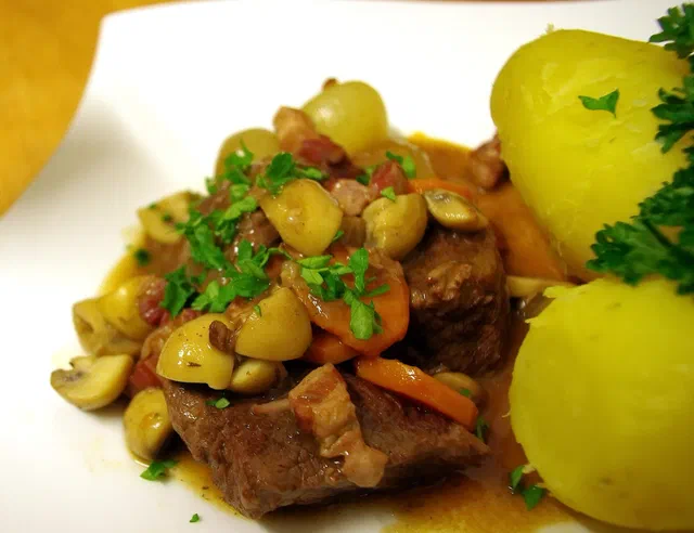 Bœuf bourguignon dans une assiette blanche : morceaux de bœuf en sauce brune, lardons, champignons, carottes et oignons grelots, avec deux pommes de terre vapeur