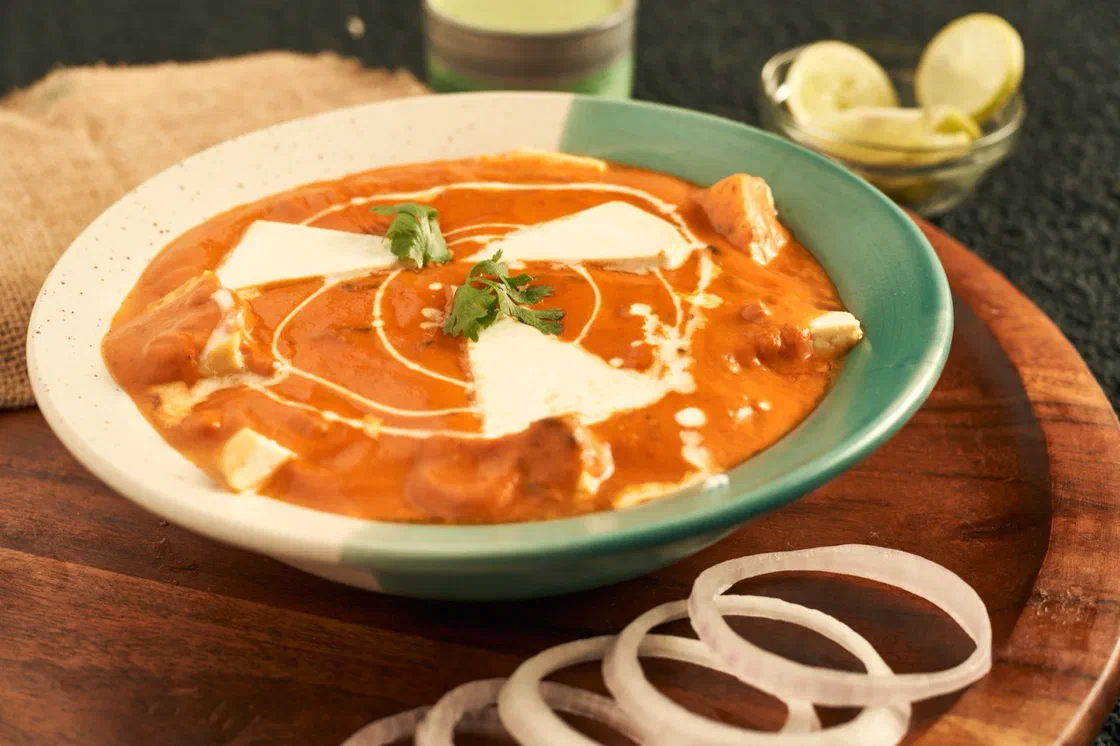 Une assiette creuse de butter chicken à la sauce orangée, avec des morceaux de poulet, un filet de crème et des feuilles de coriandre, posée sur une planche en bois à côté de rondelles d'oignon