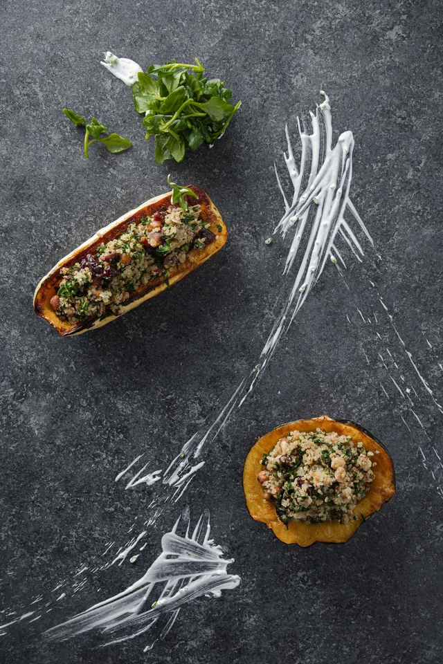 Une demi-courge longue et une tranche de courge ronde garnies d'une farce aux herbes et aux graines, sur une table en ardoise grise avec un trait de sauce blanche