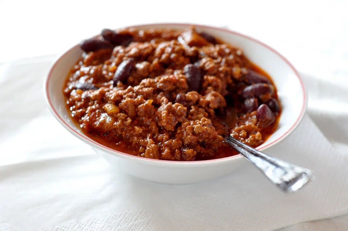 Un bol blanc de chili con carne avec de la viande hachée et des haricots rouges, une cuillère posée dans le bol