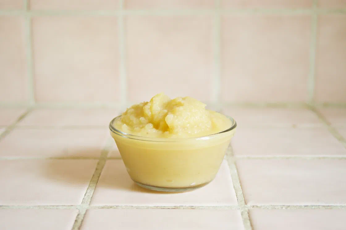 Un petit bol en verre rempli de compote de pommes jaune pâle, posé sur un carrelage blanc