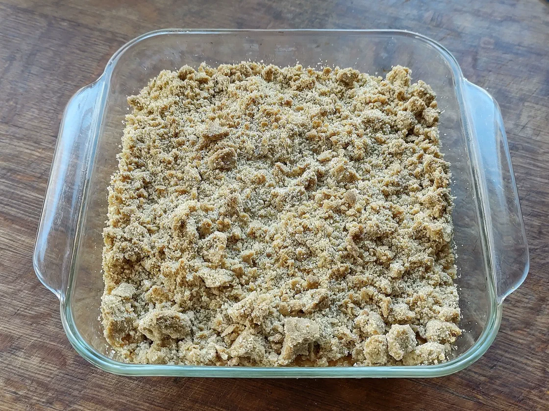 Un crumble aux pommes cuit dans un plat carré en verre, couvert d'une pâte sablée en grosses miettes