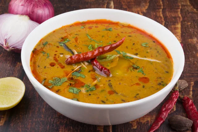 Un bol blanc de dal jaune orangé garni de piments rouges séchés et de coriandre, avec un oignon rouge et un citron à côté