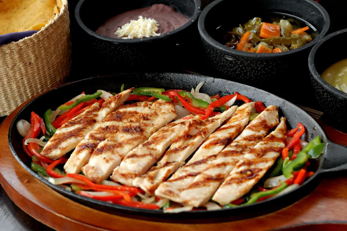 Des lanières de blanc de poulet grillé posées sur des poivrons rouges et verts et de l'oignon dans une poêle en fonte, avec des tortillas, des haricots et des sauces autour