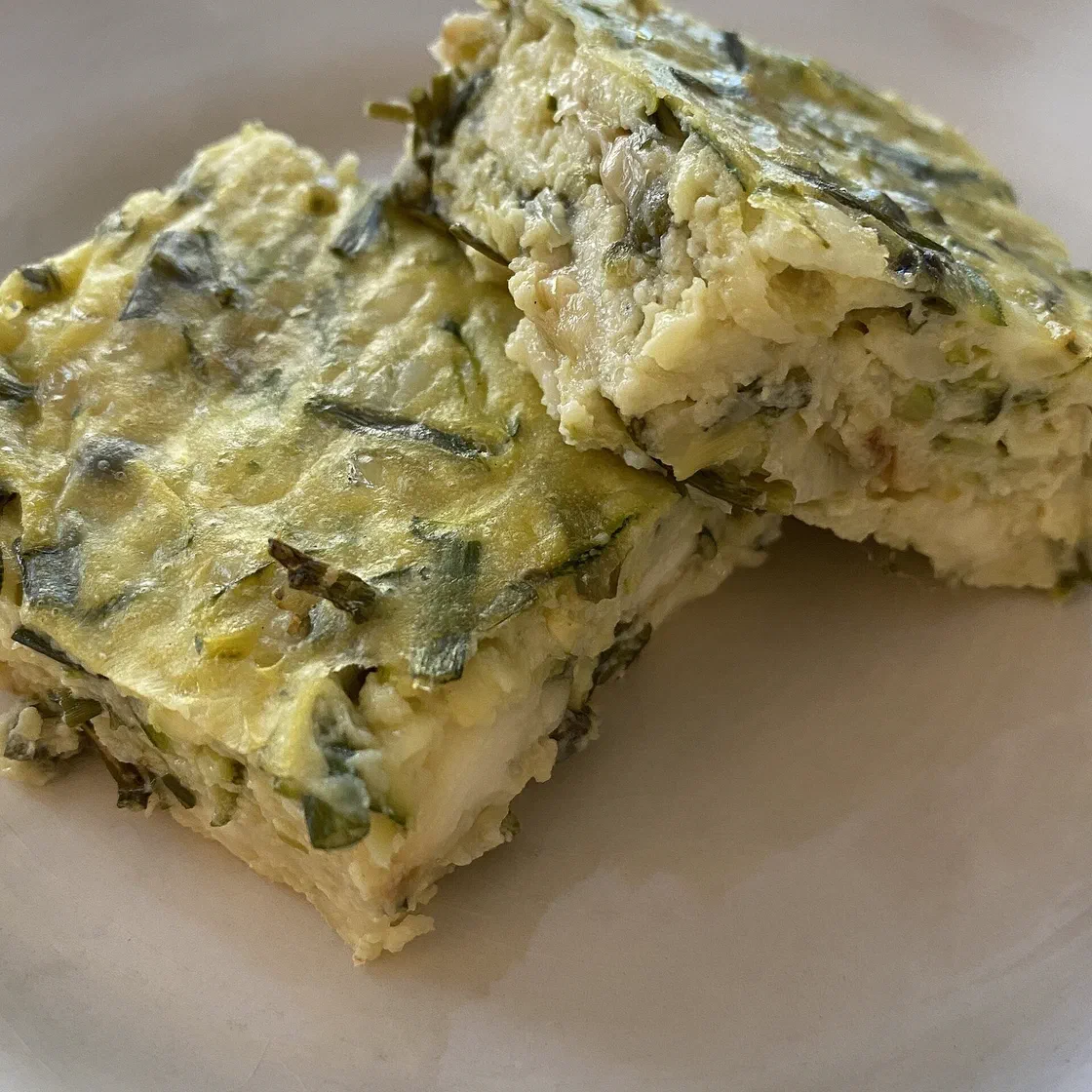 Deux parts carrées de flan de courgettes, jaune pâle et parsemées de lamelles de courgette et d'herbes, posées l'une contre l'autre sur une assiette claire