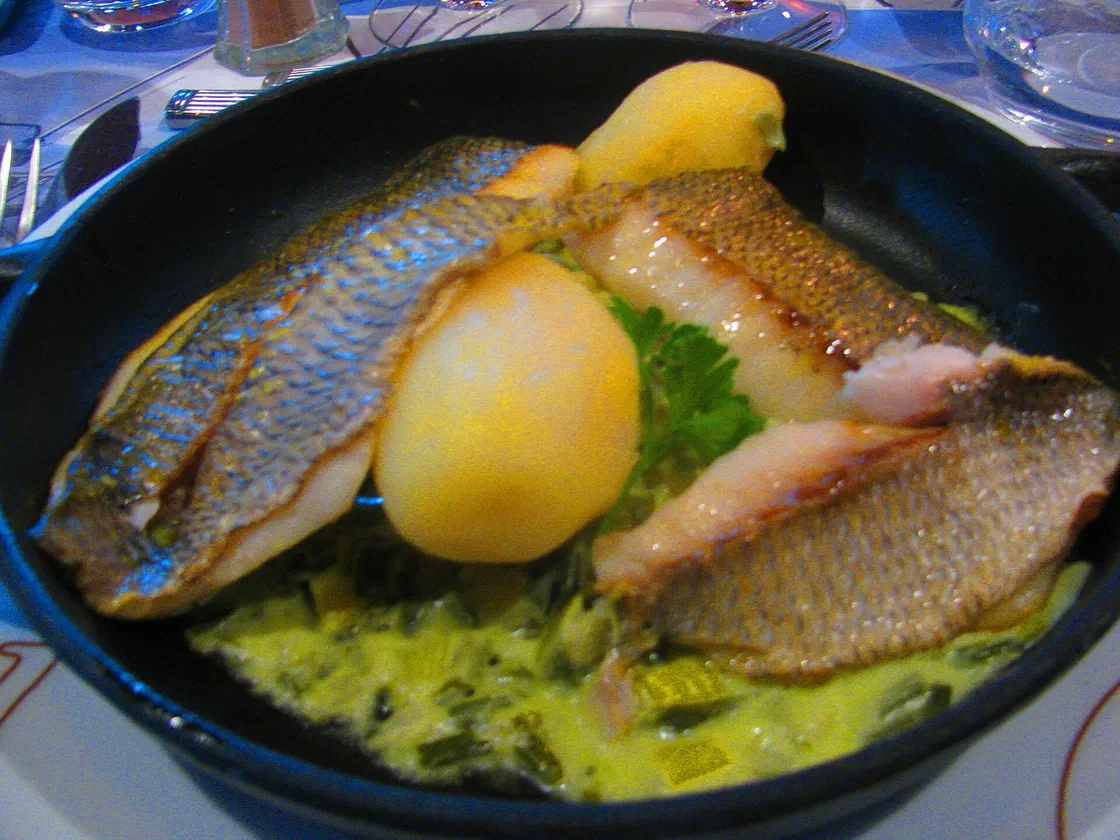 Deux filets de sar à la peau dorée posés sur une fondue de poireaux à la crème, avec deux pommes de terre, dans une poêle noire
