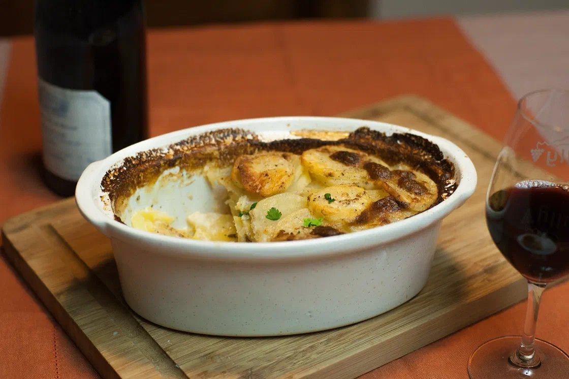 Un gratin dauphinois doré dans un plat ovale blanc posé sur une planche en bois, avec une bouteille et un verre de vin rouge