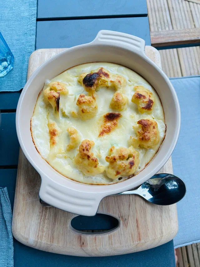 Un gratin de chou-fleur dans un plat rond blanc à anses, fleurettes dorées dépassant d'une sauce béchamel gratinée, posé sur une planche en bois