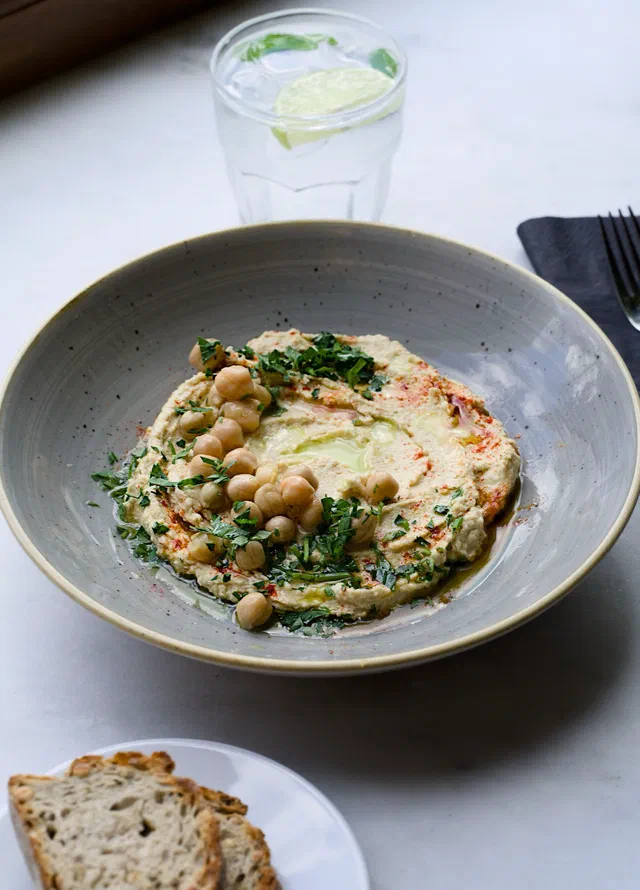 Une assiette creuse de houmous étalé, arrosé d'huile d'olive et de paprika, garni de pois chiches entiers et de persil haché, avec du pain et un verre d'eau citronnée