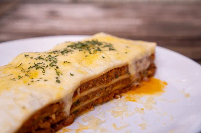 Une part de lasagnes à la bolognaise sur une assiette blanche, nappée de fromage fondu et d'herbes, avec les couches de pâte et de viande visibles sur la tranche