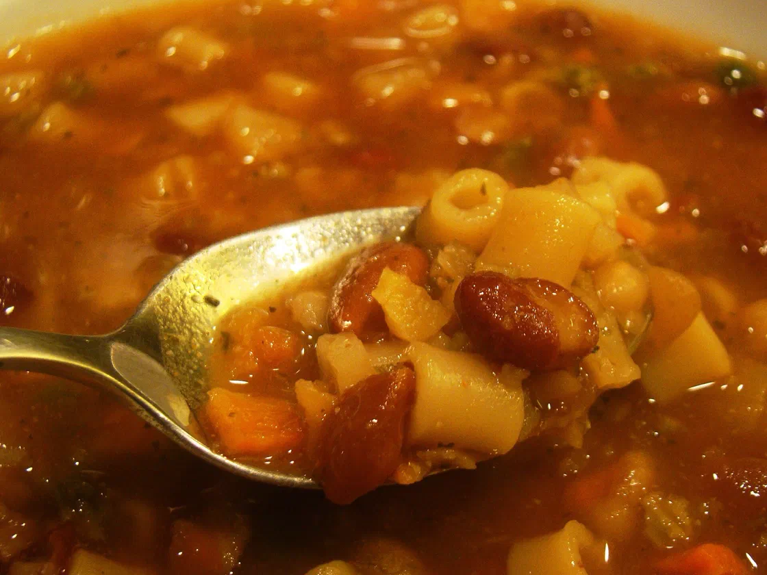 Gros plan sur une cuillère de minestrone remplie de petites pâtes tubes, de haricots rouges et de dés de carotte, au-dessus d'un bouillon tomaté