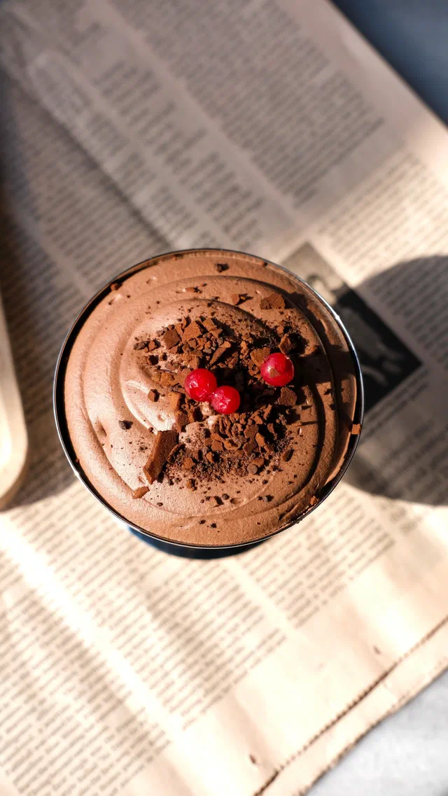 Une verrine de mousse au chocolat vue de dessus, saupoudrée de cacao et garnie de groseilles, posée sur un journal