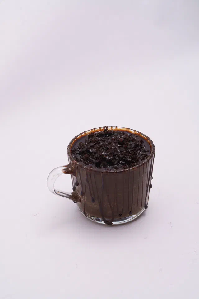 Un mug cake au chocolat dans une tasse en verre, le dessus couvert de copeaux de chocolat, sur fond blanc