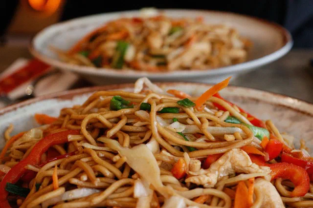 Gros plan sur une assiette de nouilles aux œufs sautées avec des morceaux de poulet, du chou, de la carotte, du poivron rouge et de l'oignon nouveau, une seconde assiette en arrière-plan