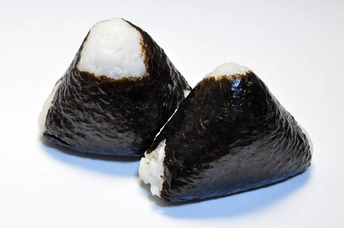 Deux onigiri triangulaires en riz blanc enveloppés d'une feuille de nori, posés sur un fond blanc