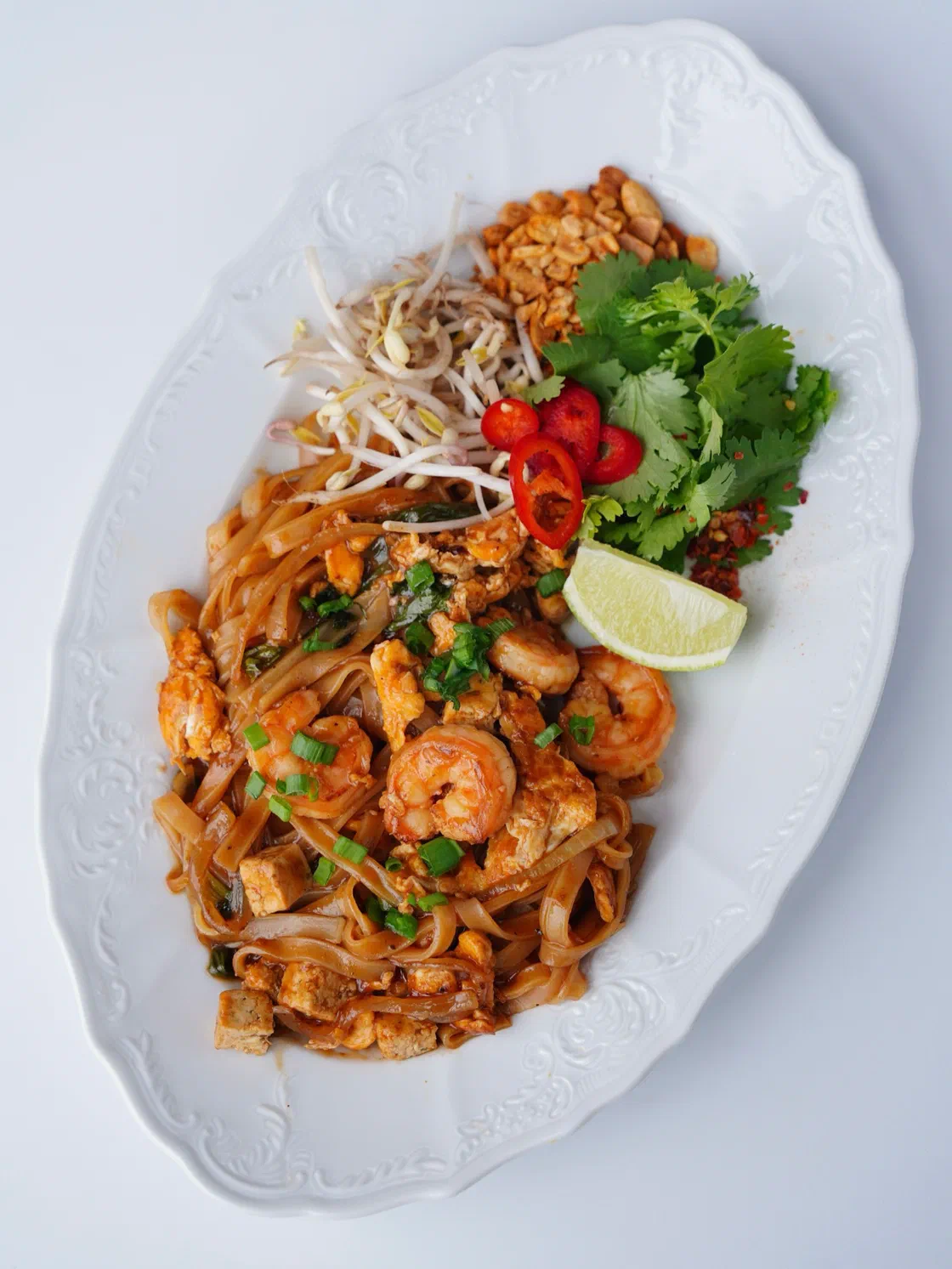 Un pad thaï aux crevettes sur un plat ovale blanc, avec pousses de soja, coriandre, piment rouge et un quartier de citron vert