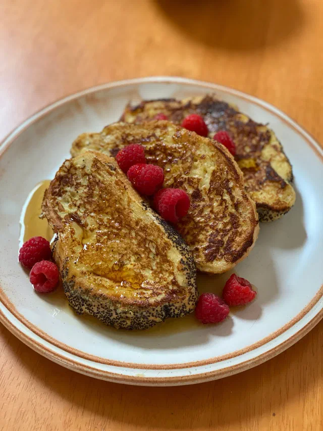Trois tranches de pain perdu bien dorées sur une assiette à bord brun, arrosées de sirop et parsemées de framboises