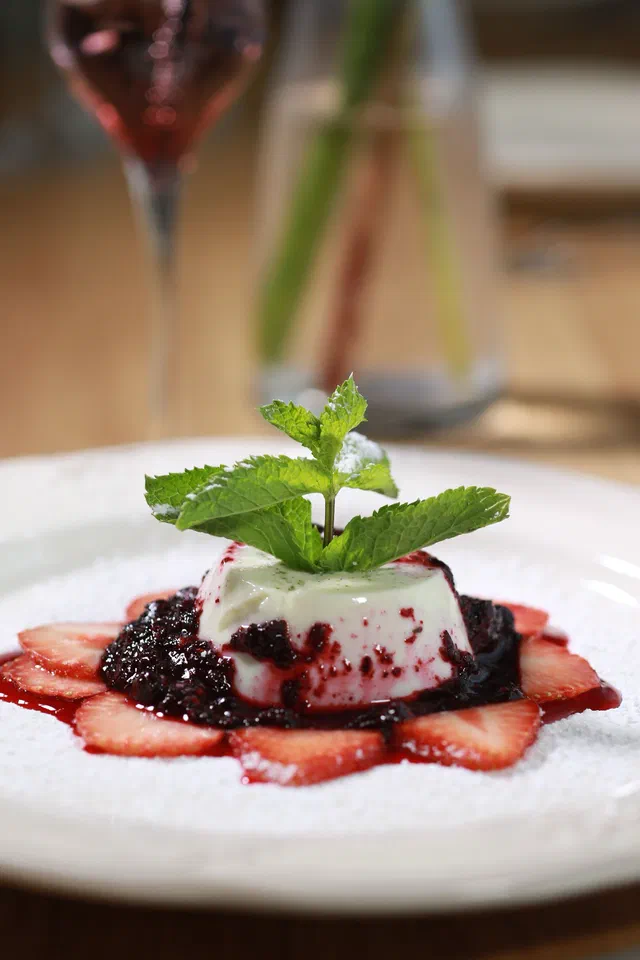 Une panna cotta démoulée nappée de coulis de fruits rouges, avec des tranches de fraise et des feuilles de menthe sur une assiette blanche
