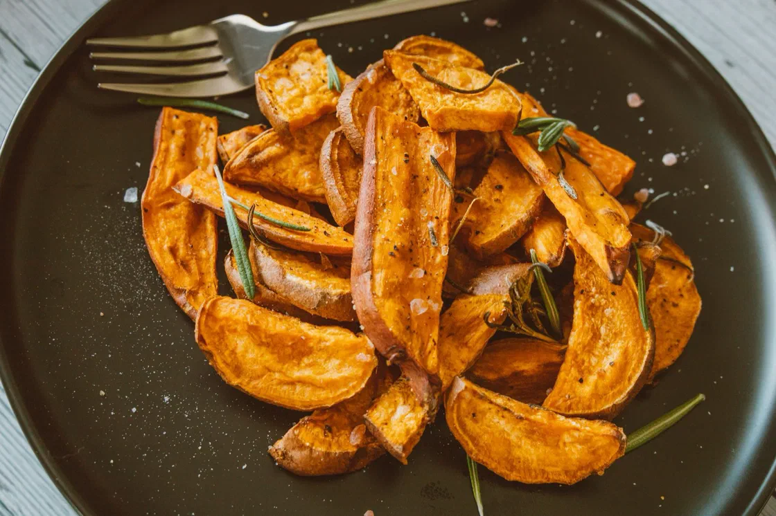 Quartiers de patate douce rôtis au four avec leur peau, parsemés de brins de romarin, dans une assiette noire avec une fourchette