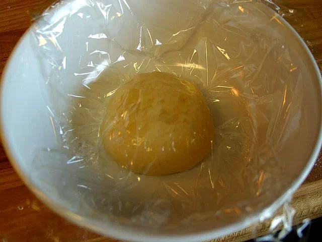 Une boule de pâte à pizza levée dans un saladier blanc couvert de film alimentaire