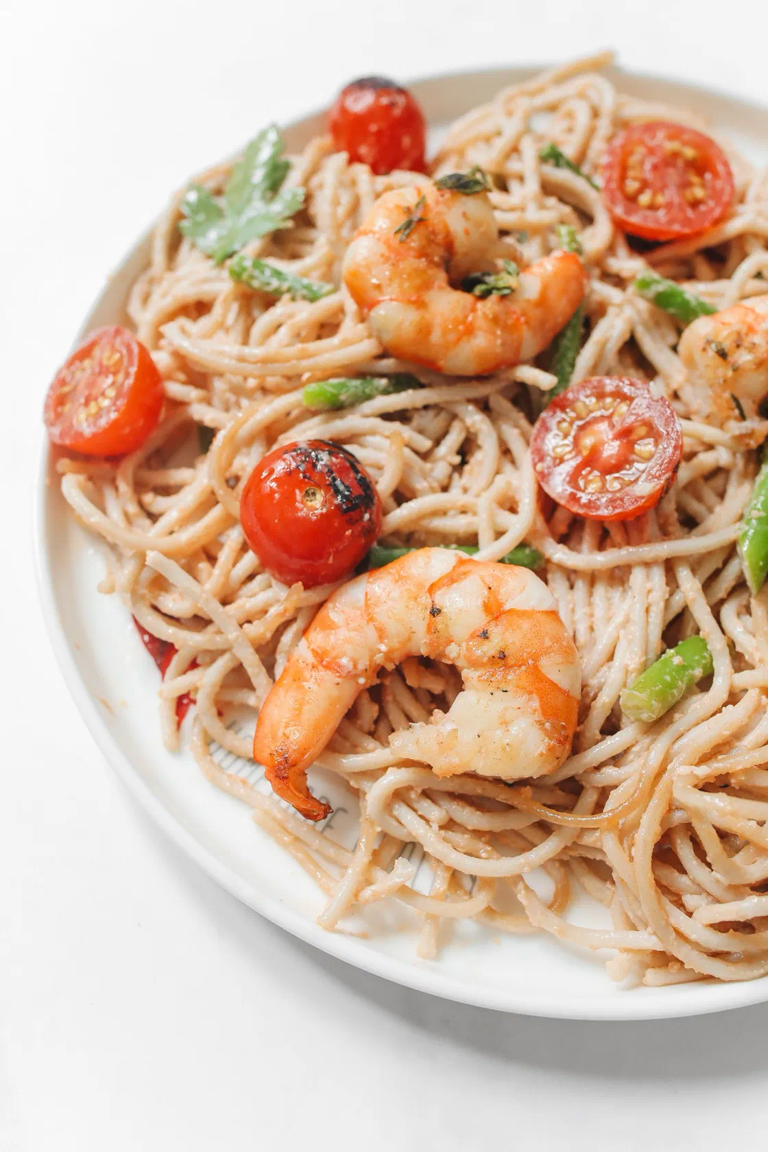Une assiette blanche de spaghetti aux crevettes dorées, avec des tomates cerises entières et coupées, des feuilles de persil et quelques tronçons d'asperge verte