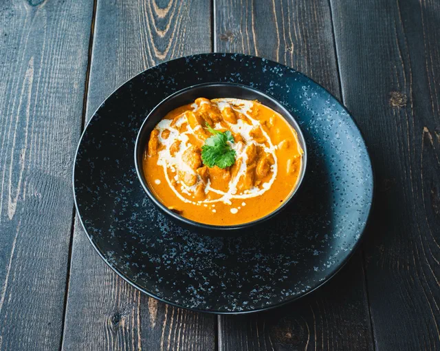 Un bol noir de curry de poulet en sauce orangée, nappé d'un filet de crème de coco et garni d'une feuille de coriandre, posé sur une table en bois sombre