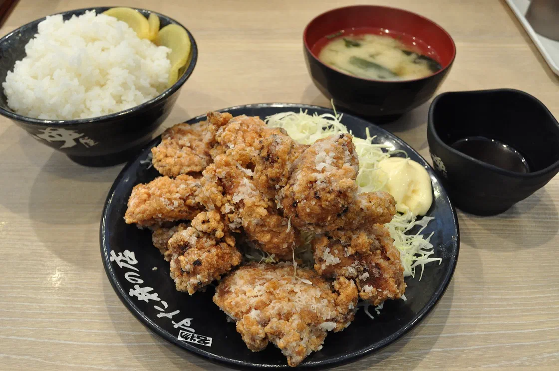 Une assiette noire de morceaux de poulet karaage frits et dorés sur un lit de chou émincé, avec un bol de riz blanc et un bol de soupe miso à l'arrière