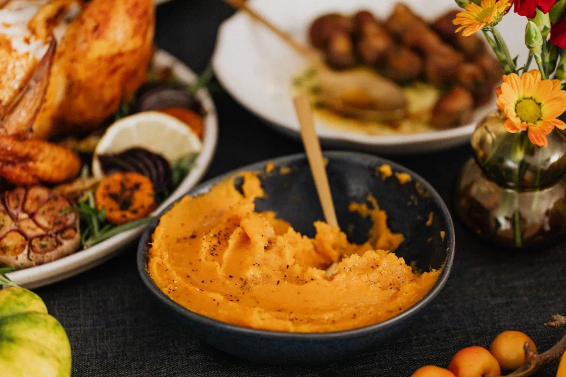 Purée de patate douce orange poivrée dans un bol gris avec une cuillère dorée, devant un poulet rôti, des légumes rôtis et un bouquet de fleurs jaunes