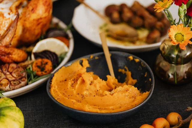 Purée de patate douce orange poivrée dans un bol gris avec une cuillère dorée, devant un poulet rôti, des légumes rôtis et un bouquet de fleurs jaunes