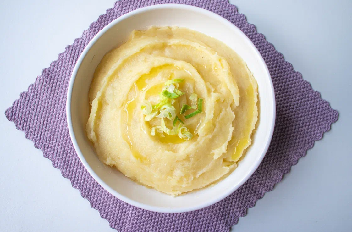 Un bol blanc de purée de pommes de terre lisse et jaune pâle, avec un filet d'huile et de la ciboule émincée, sur un set violet