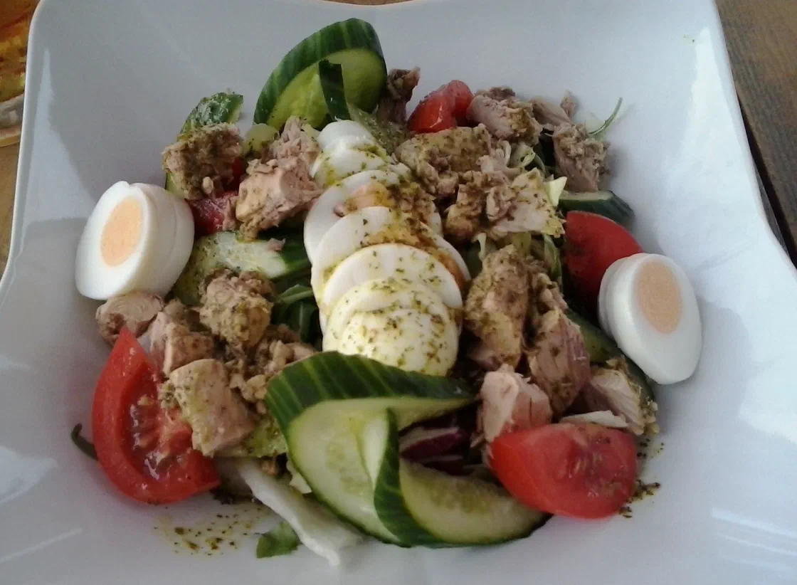 Salade de thon en morceaux avec des demi-œufs durs, des quartiers de tomate, des rondelles de concombre et des tranches de mozzarella sur un lit de laitue, dans une assiette creuse blanche