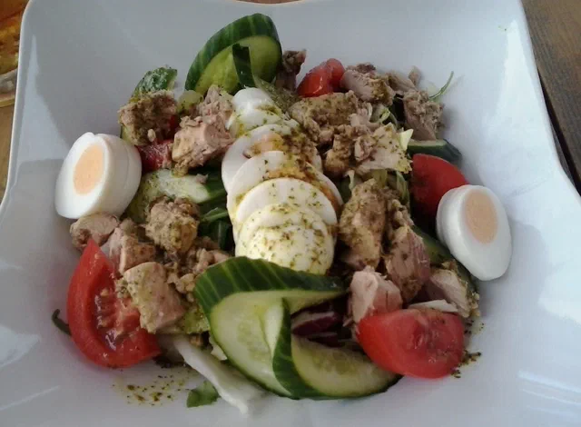 Salade de thon en morceaux avec des demi-œufs durs, des quartiers de tomate, des rondelles de concombre et des tranches de mozzarella sur un lit de laitue, dans une assiette creuse blanche