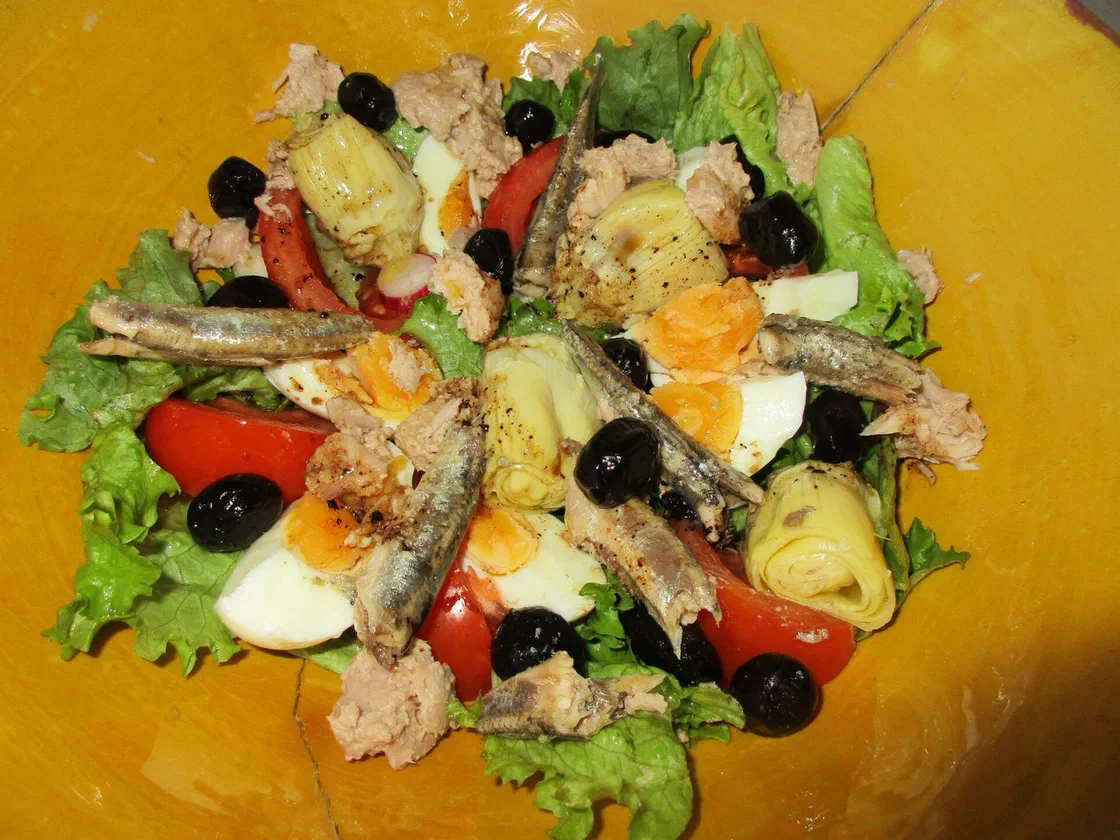 Une salade niçoise dans un plat jaune : feuilles de laitue, quartiers de tomate, œufs durs, thon, sardines ou anchois, cœurs d'artichaut et olives noires