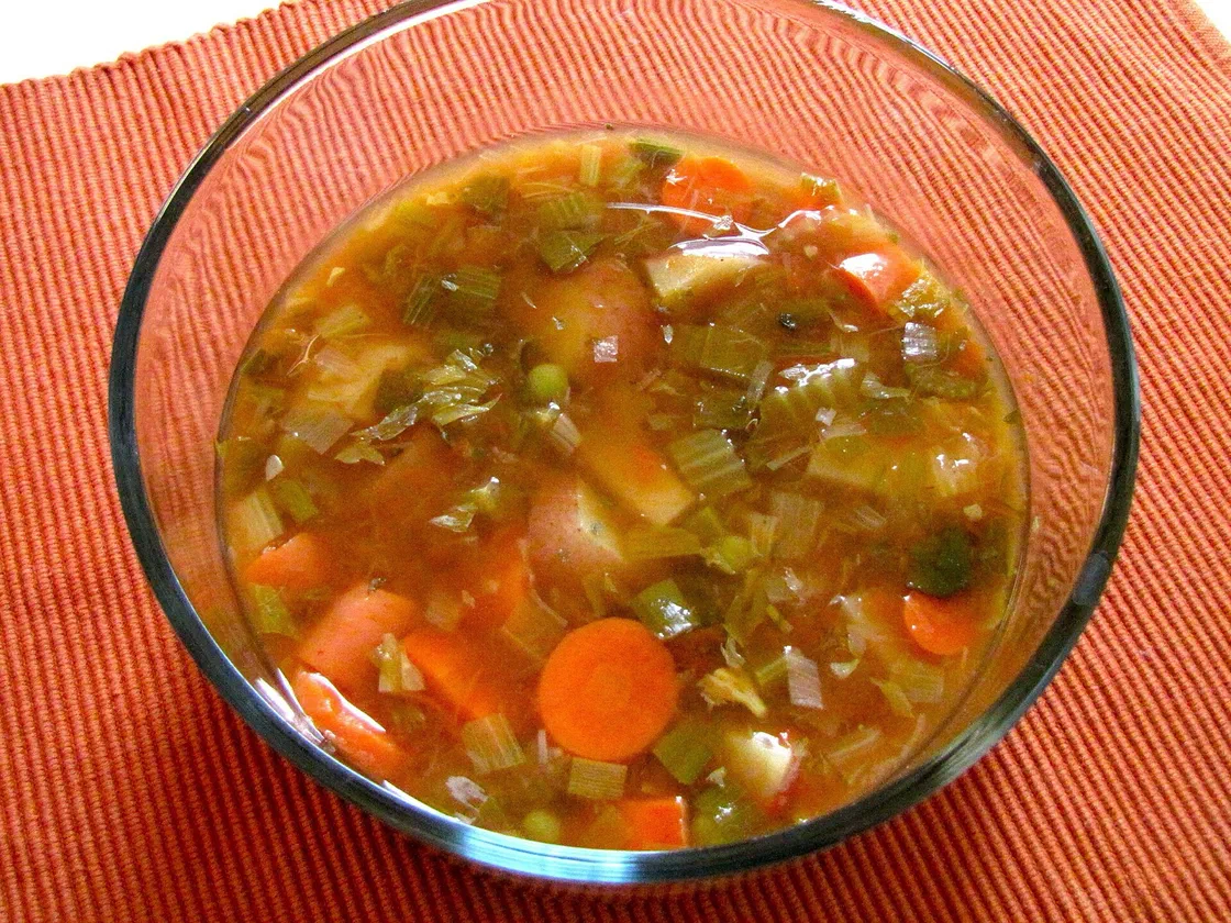 Saladier en verre rempli de soupe de légumes en morceaux, rondelles de carotte, pommes de terre, céleri et petits pois dans un bouillon tomaté, posé sur un set orange