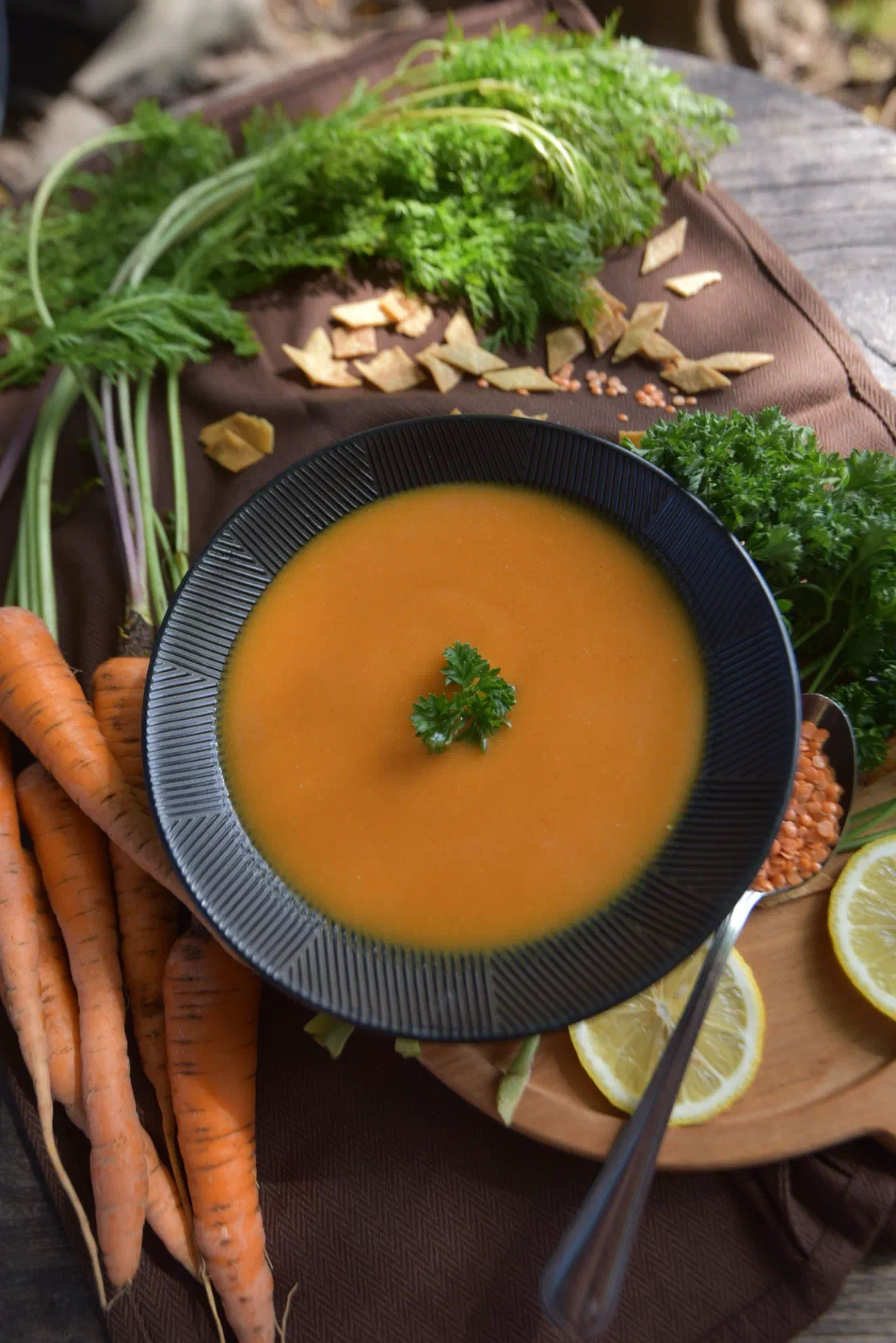 Une assiette creuse de soupe de légumes mixée orange, une feuille de persil au centre, entourée de carottes crues et de fanes