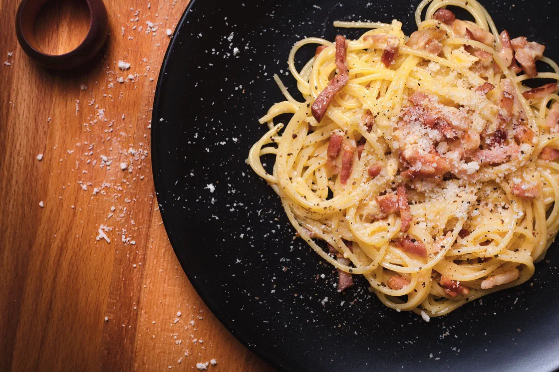 Des spaghetti carbonara avec des dés de guanciale grillés et du fromage râpé dans une assiette noire posée sur une planche en bois