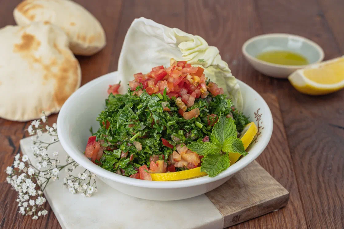 Un bol blanc de taboulé où domine le persil haché, avec des dés de tomate, de la menthe et un quartier de citron, à côté de pains pita et d'une coupelle d'huile d'olive