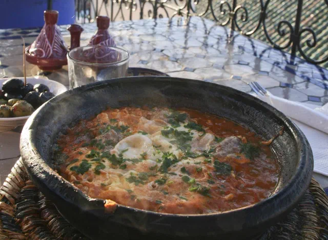 Tajine en terre noire rempli de sauce tomate, de boulettes de viande et d'œufs cuits, parsemé de persil, sur une table en terrasse