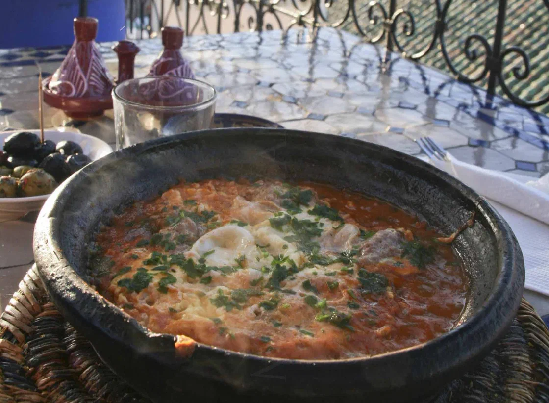 Tajine en terre noire rempli de sauce tomate, de boulettes de viande et d'œufs cuits, parsemé de persil, sur une table en terrasse