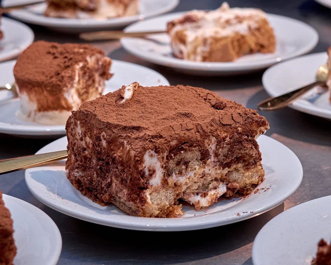 Une part de tiramisu couverte de cacao sur une assiette blanche, d'autres parts servies en arrière-plan