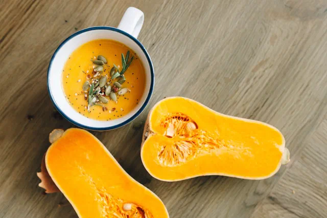 Un velouté de butternut orange dans une tasse, garni de graines et de romarin, à côté d'une courge butternut coupée en deux