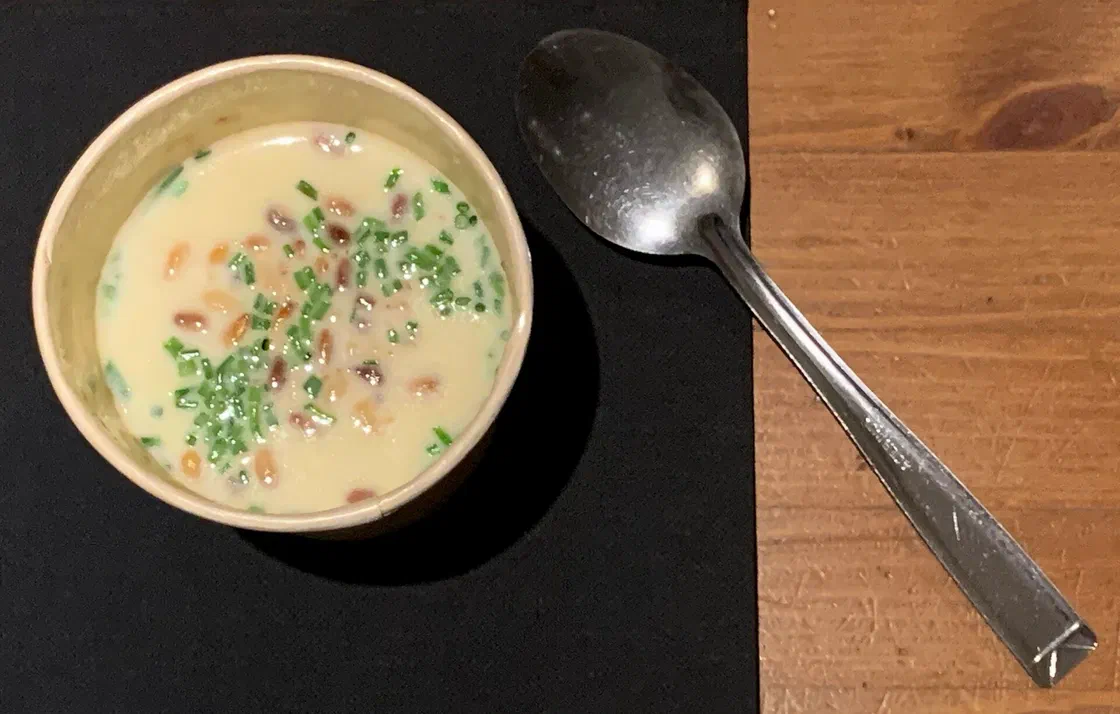 Un bol de velouté de panais crème parsemé de ciboulette ciselée et de pignons de pin, posé sur un set noir à côté d'une cuillère