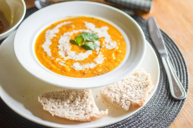 Un velouté de potiron orange dans une assiette creuse blanche, avec un filet de crème en spirale et une feuille d'herbe, deux tranches de pain à côté